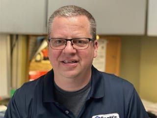 Man with glasses wearing a dark blue polo shirt smiles indoors.