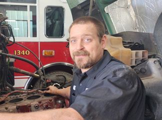 Mechanic working on a truck engine in a garage with a red fire truck in the background.