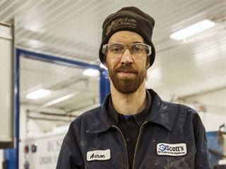 Man in work uniform, safety glasses, and beanie smiles in a garage.
