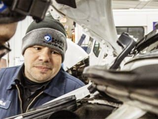 Mechanic in blue uniform and beanie, inspecting car engine, looking intently. Shop interior.