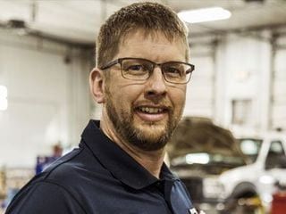 Man with glasses smiling in a garage, wearing a dark blue collared shirt.