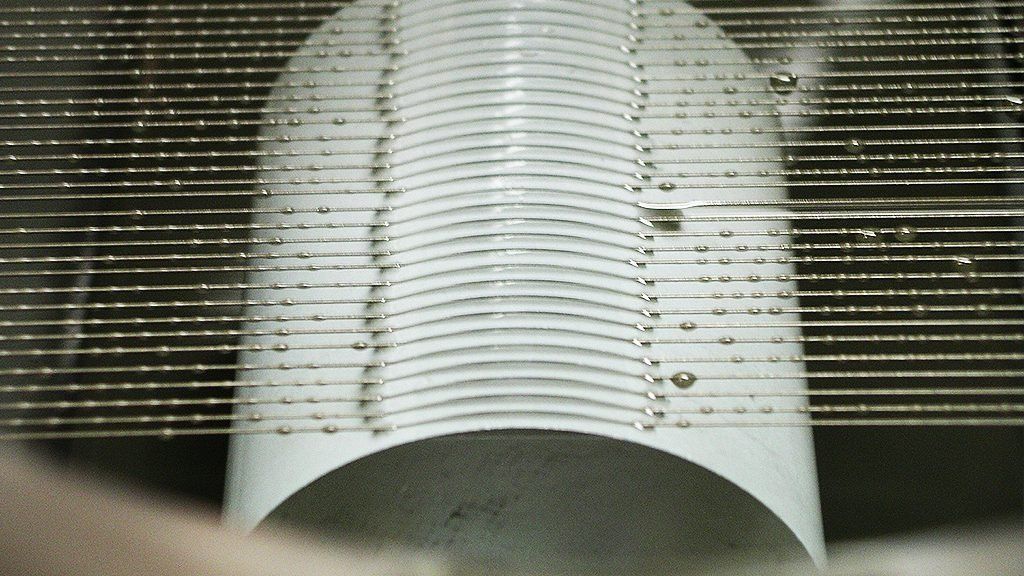 A person in a cleanroom suit and gloves is handling a silicon wafer with a shiny, reflective surface. The wafer is held over a transparent container, with another wafer inside. Computer monitors are visible in the background.