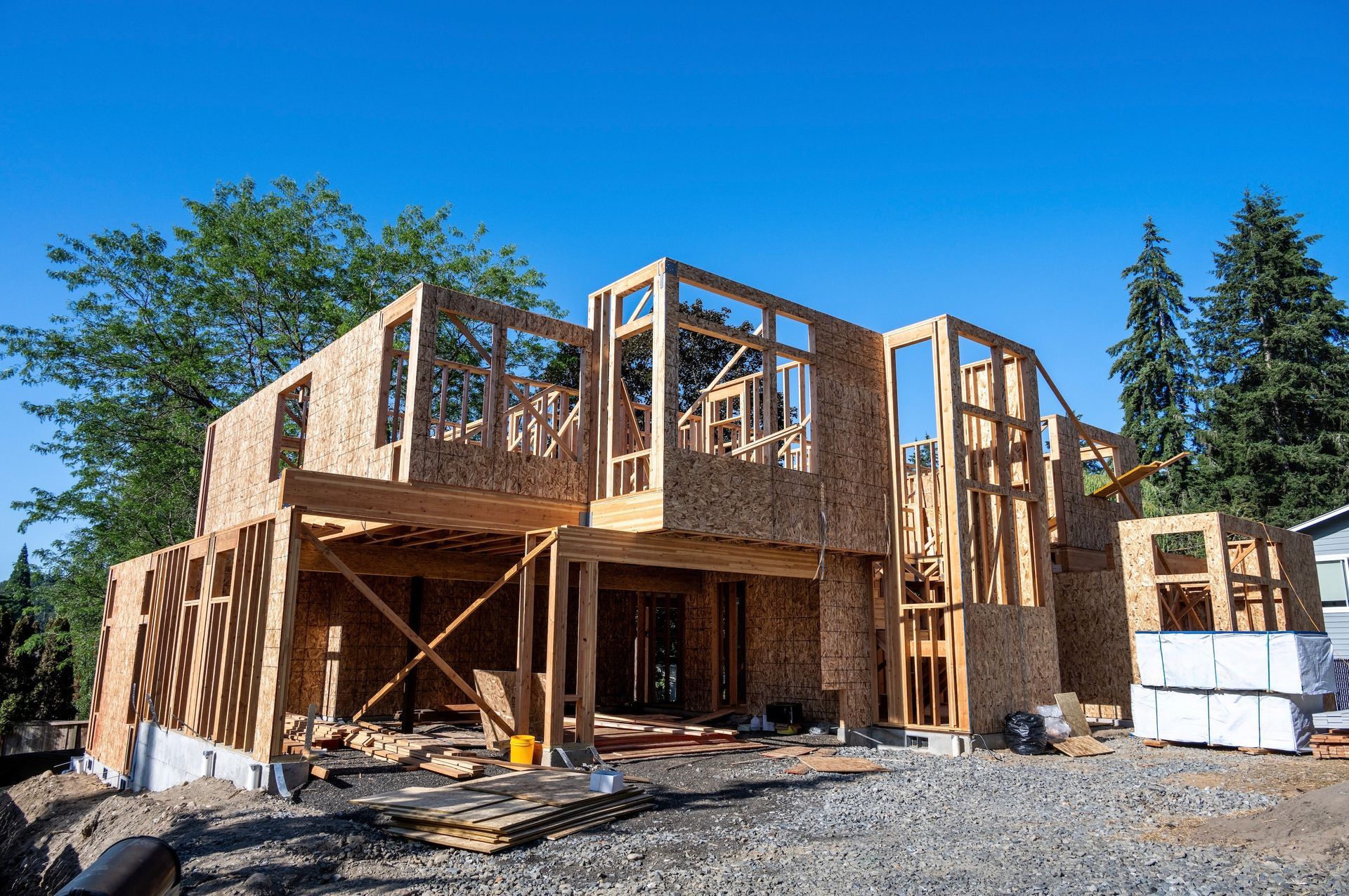 Two-story house under construction; wooden frame exposed; gravel in foreground; blue sky.
