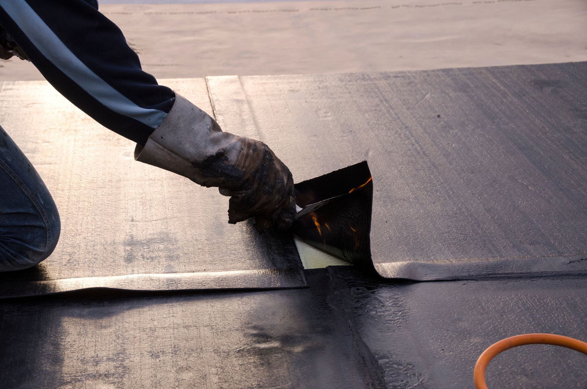 Person applying sealant to a dark, flat roofing surface, using a trowel and wearing a protective glove.