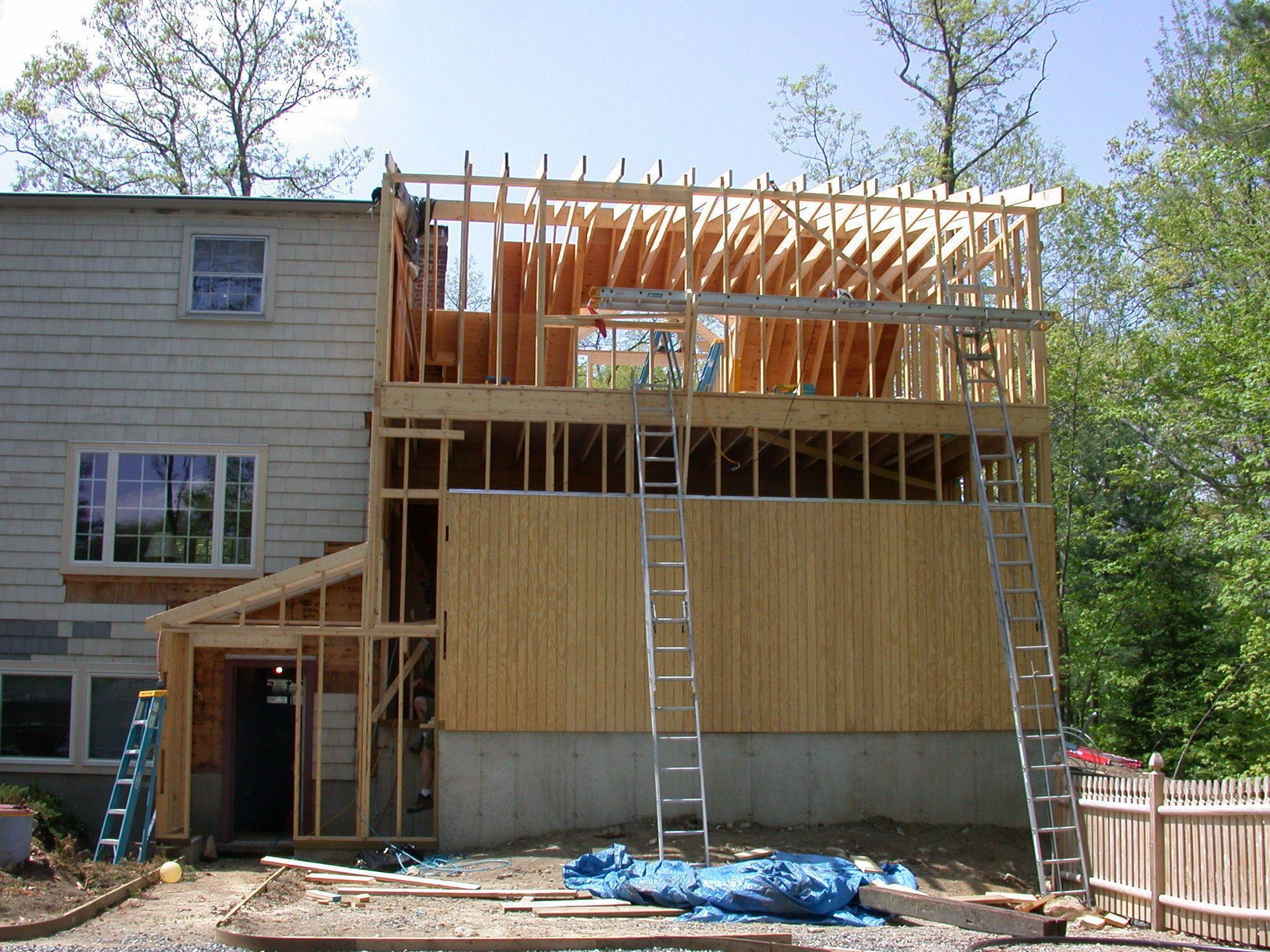 Building addition under construction, wooden frames, siding, ladders, surrounded by trees.