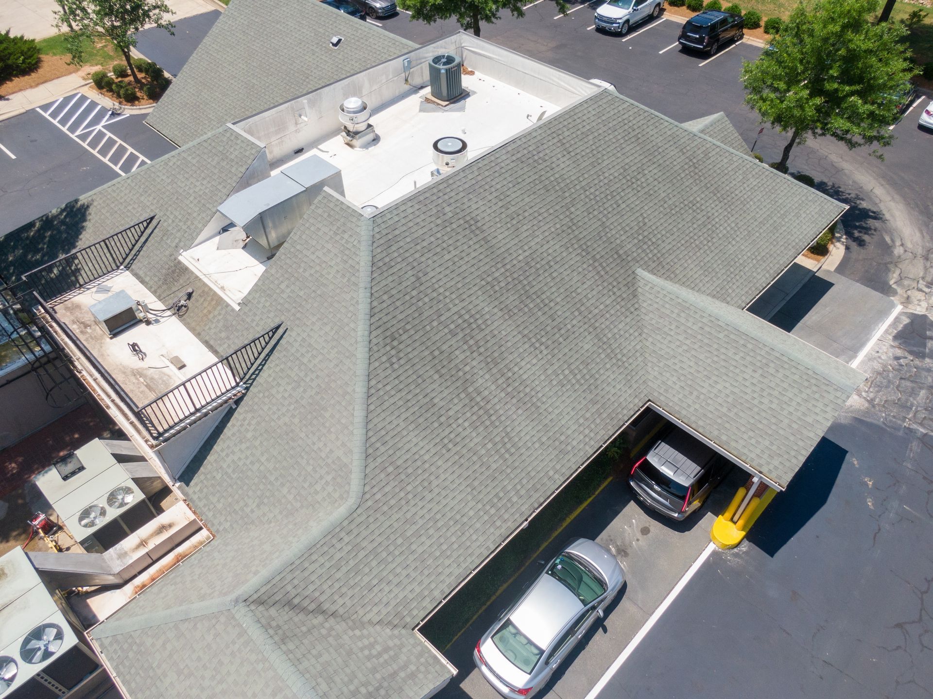 Aerial view of a building with a gray shingle roof, surrounded by a parking lot and trees. Cars are parked nearby.
