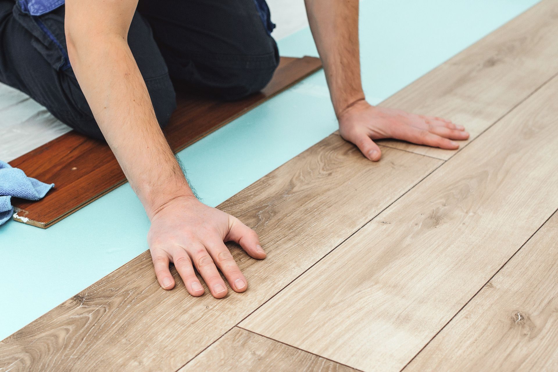 Person kneeling, installing light brown laminate flooring on a blue underlayment.