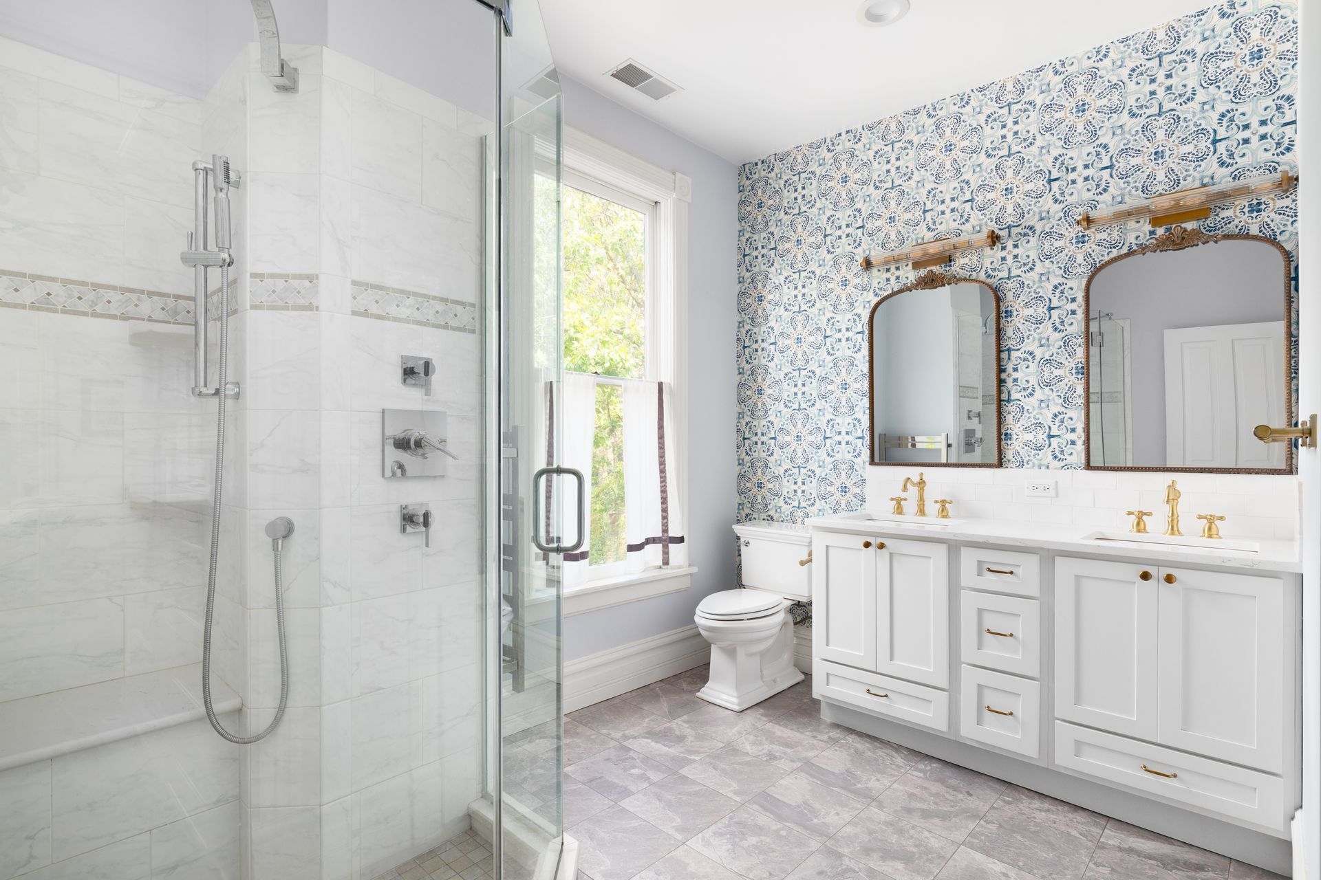 Bathroom with marble shower, double vanity, blue patterned wallpaper, and gray tiled floor.
