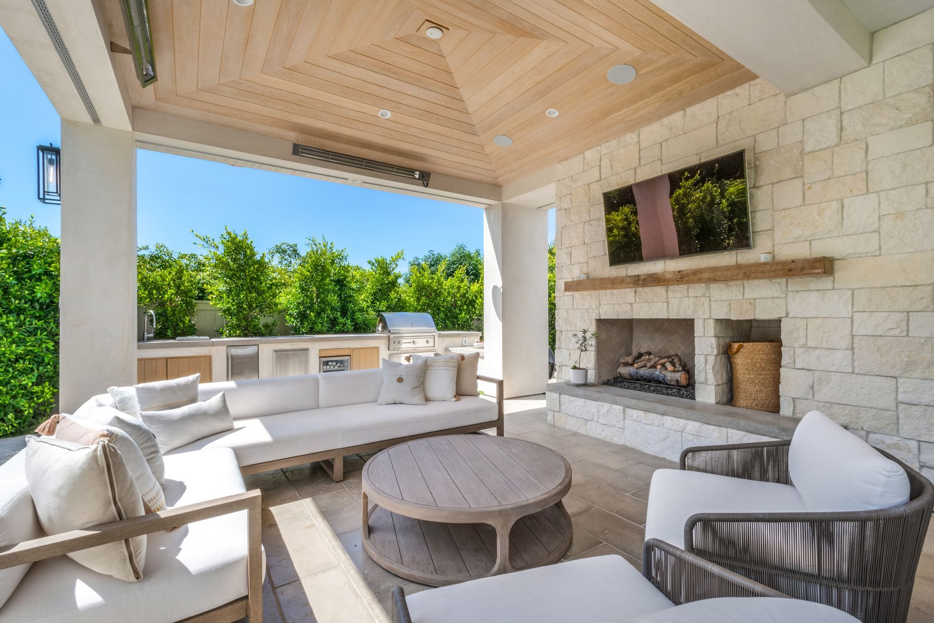 Outdoor patio with seating, fireplace, grill, and TV; neutral tones and greenery.