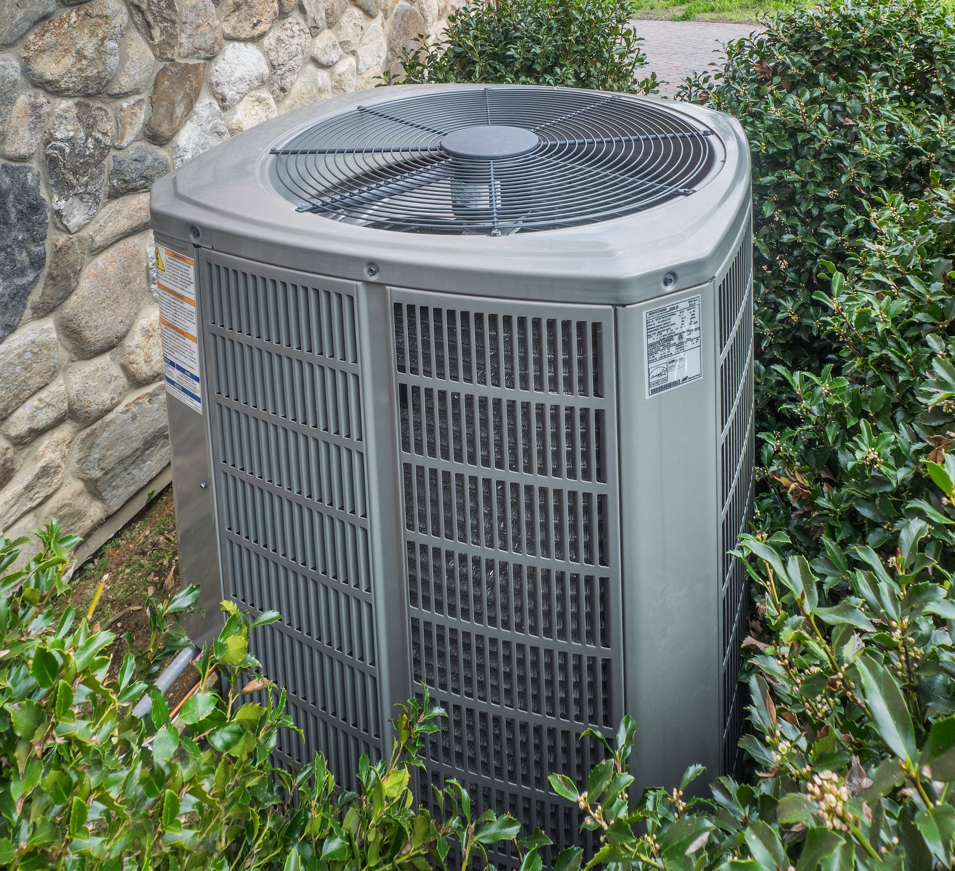 Outdoor air conditioning unit, gray, surrounded by green shrubs, near a stone wall.