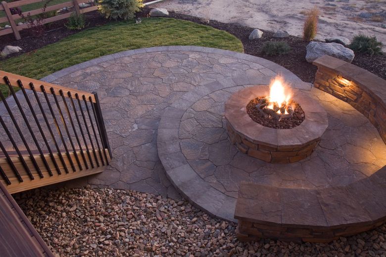 Circular stone patio with fire pit and built-in seating, near a wooden deck and landscaping.