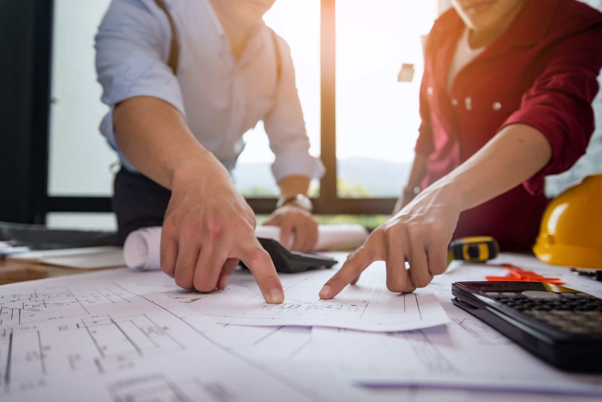 Two people pointing at architectural blueprints on a desk in a bright office.