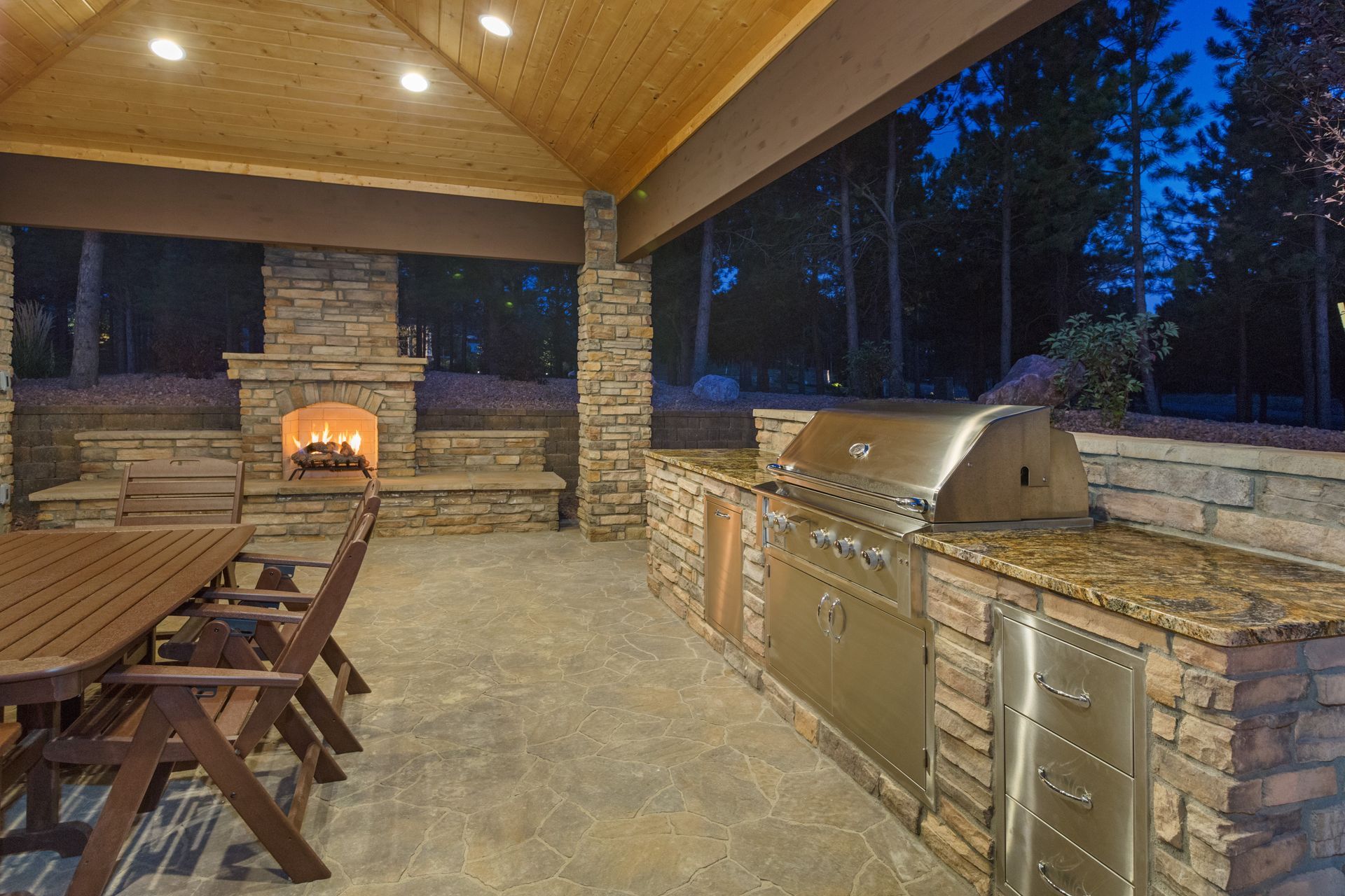 Outdoor kitchen and fireplace area at dusk with a grill, countertop, and seating.