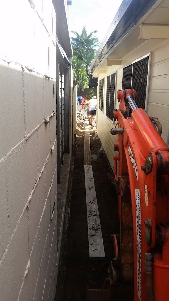 A Red Excavator Is Digging A Trench Between Two Buildings — Absolute Dingo Services in White Rock, QLD