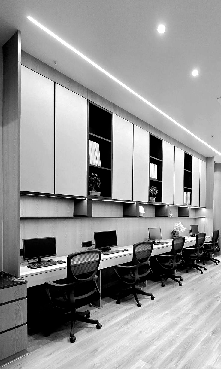 Black and white office workspace with desks, chairs, computers, and overhead storage cabinets.