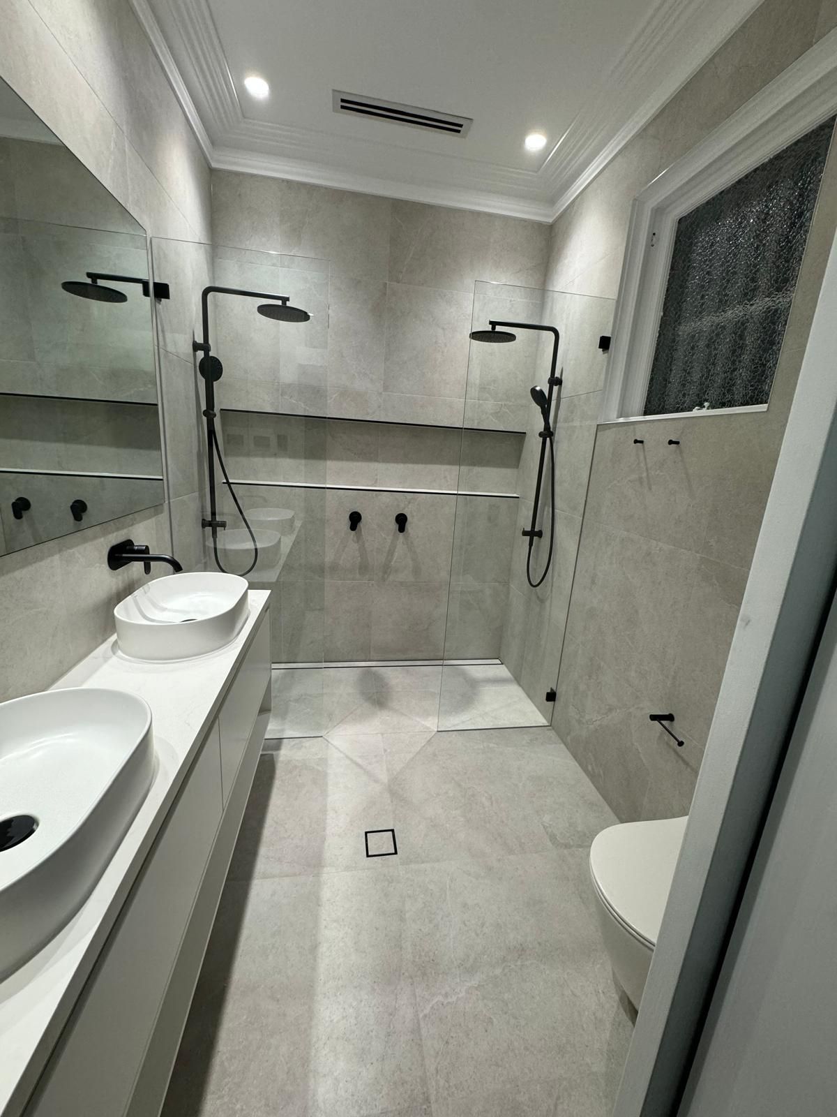 Modern bathroom with dual shower heads, white sinks, toilet, and light gray tiles.