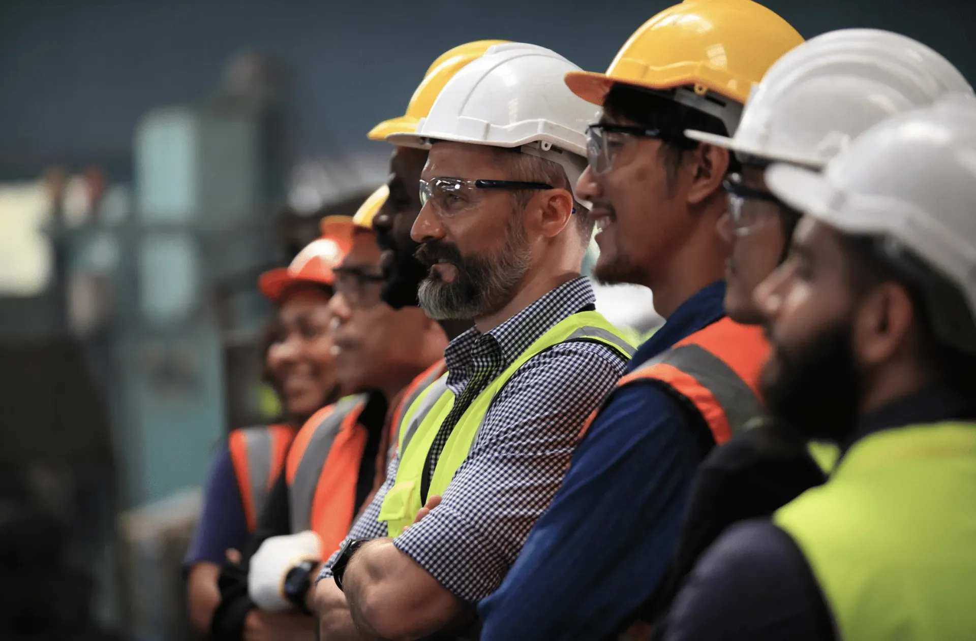 Construction workers in hard hats and vests, standing in a factory, some wearing glasses.