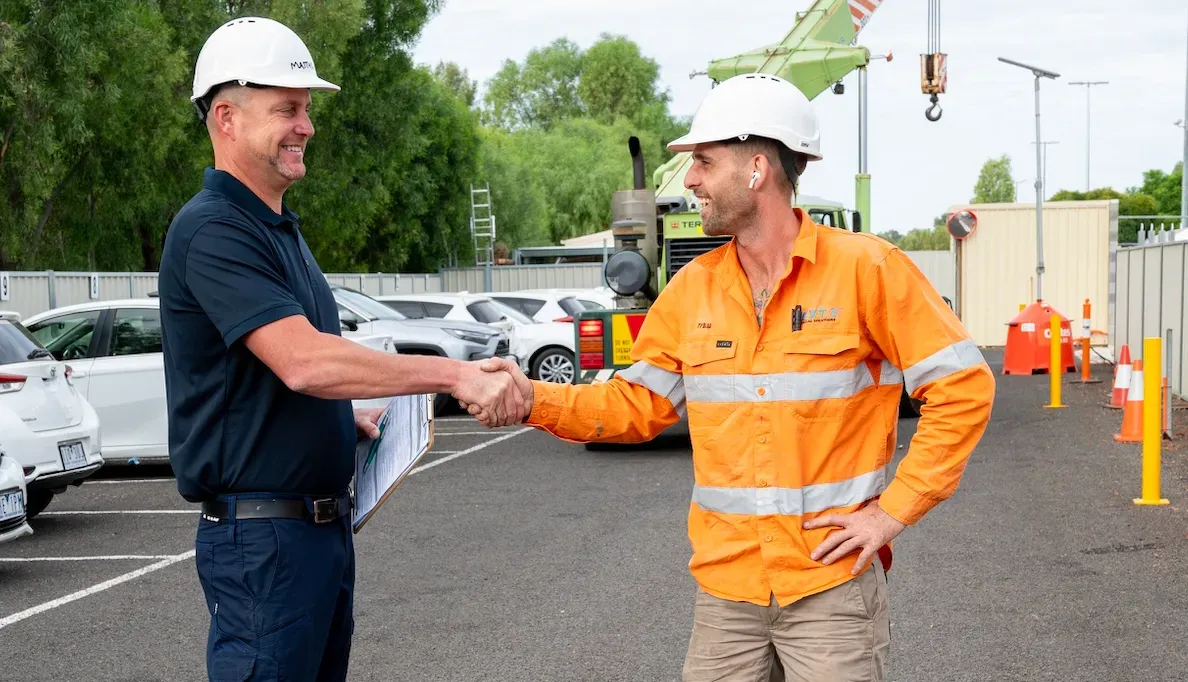 Two men in hard hats shake hands in a parking lot, a crane visible in the background.