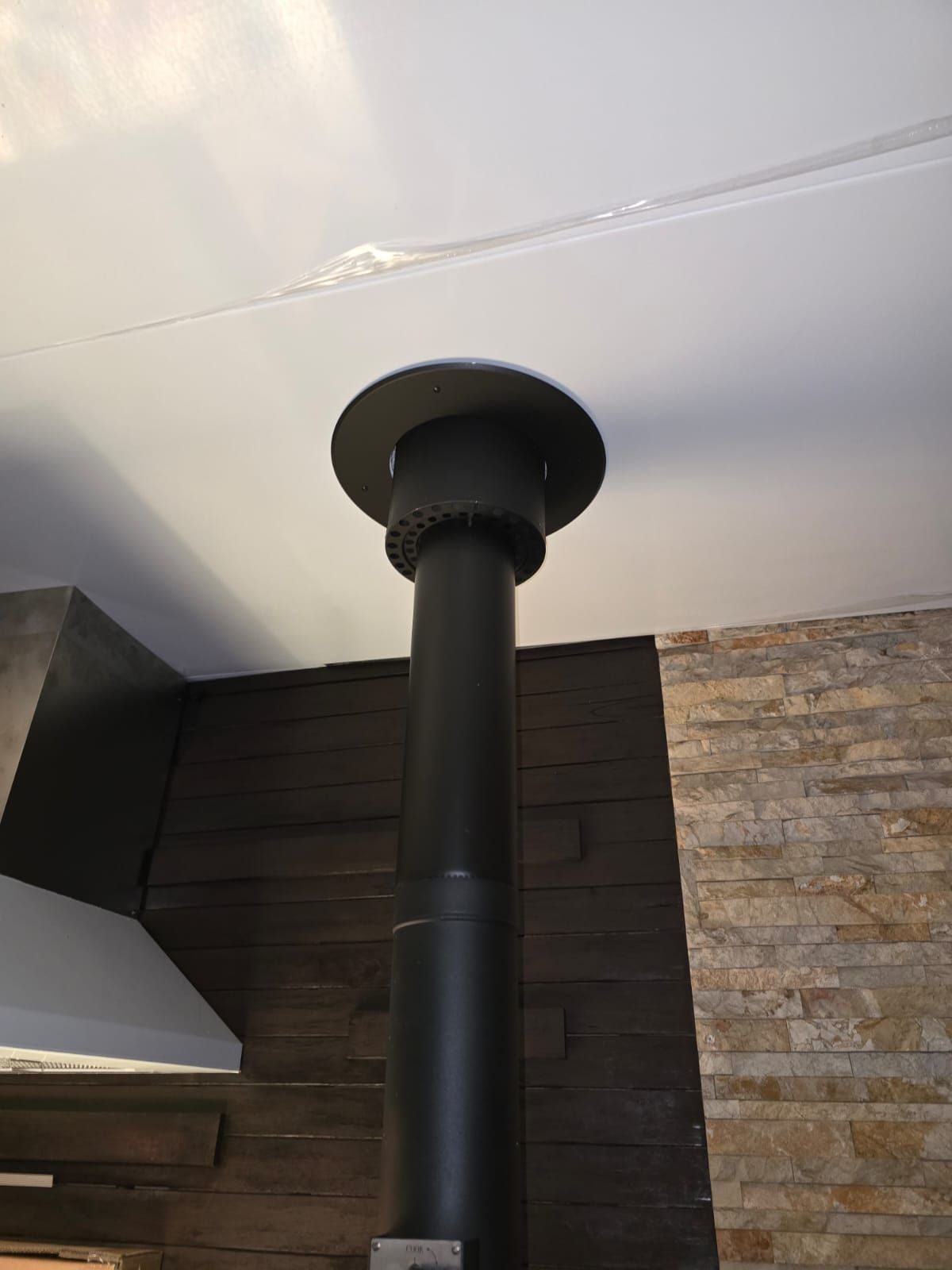 Black stovepipe extending through a white ceiling. Decorative black cap. Wood paneling and stone wall in background.