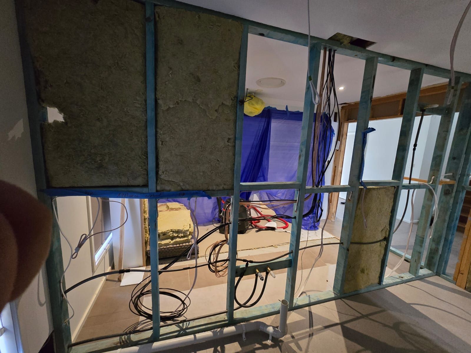 Interior wall under construction, wooden frame, insulation, wires. Blue tarp in the background.