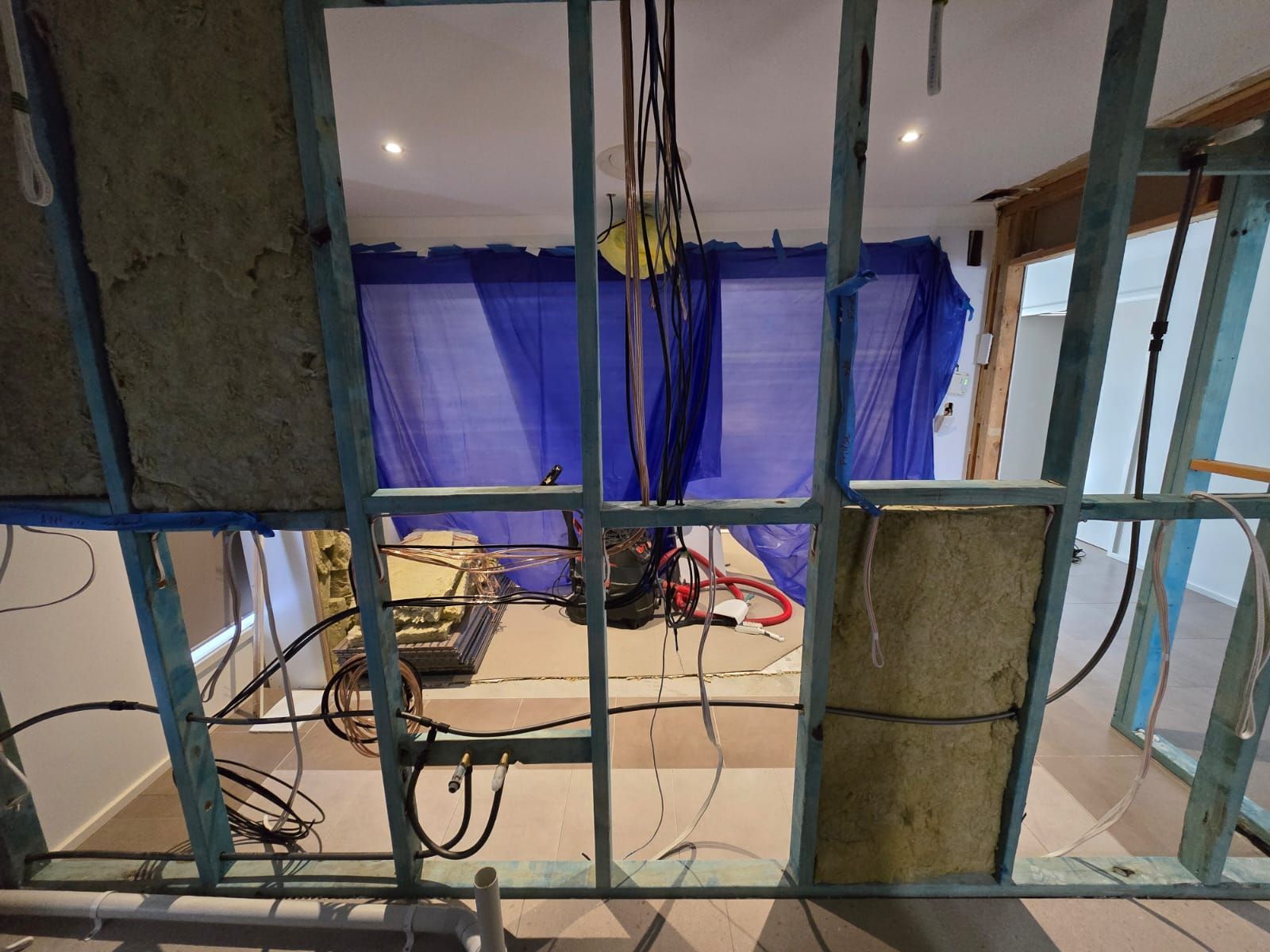 Interior construction view through framing, with blue tarps, insulation, and exposed wiring.