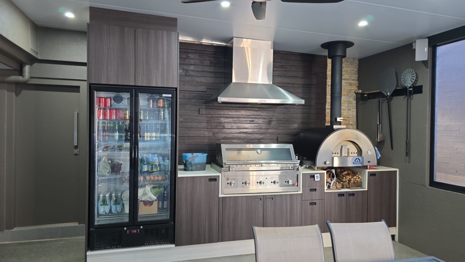 Outdoor kitchen with grill, pizza oven, refrigerator, and dark wood cabinets.