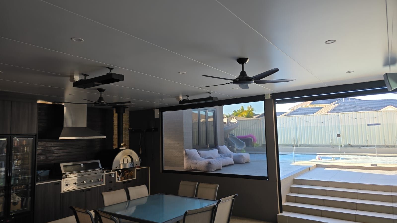 Outdoor kitchen with a projector screen displaying a backyard pool scene, under a white ceiling.
