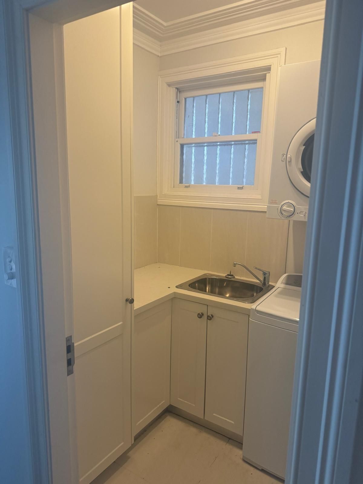 Small laundry room with white cabinets, stainless steel sink, washer, dryer, and window.