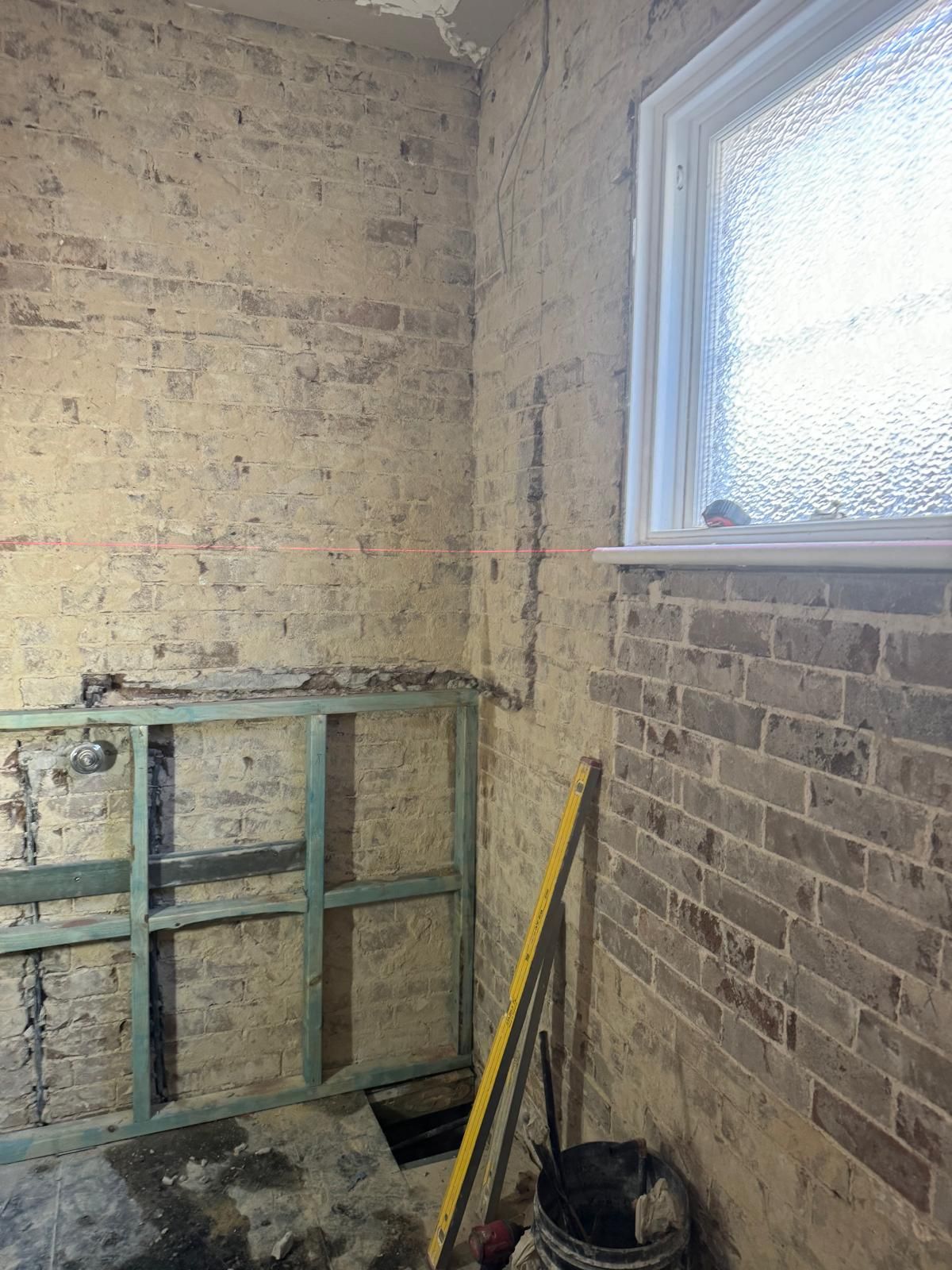 Brick walls in a room under construction, with a window and wooden frame. A level and bucket are present.