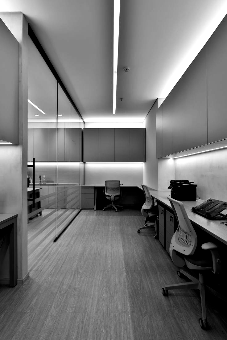 Modern office with desks, chairs, and storage, lit by long overhead and wall lights. Black and white.
