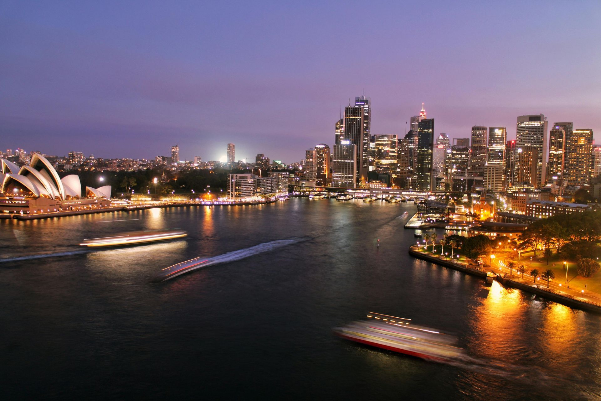 Sydney Harbour at dusk, with the Opera House on the left and city skyline in the background. Boats on the water.