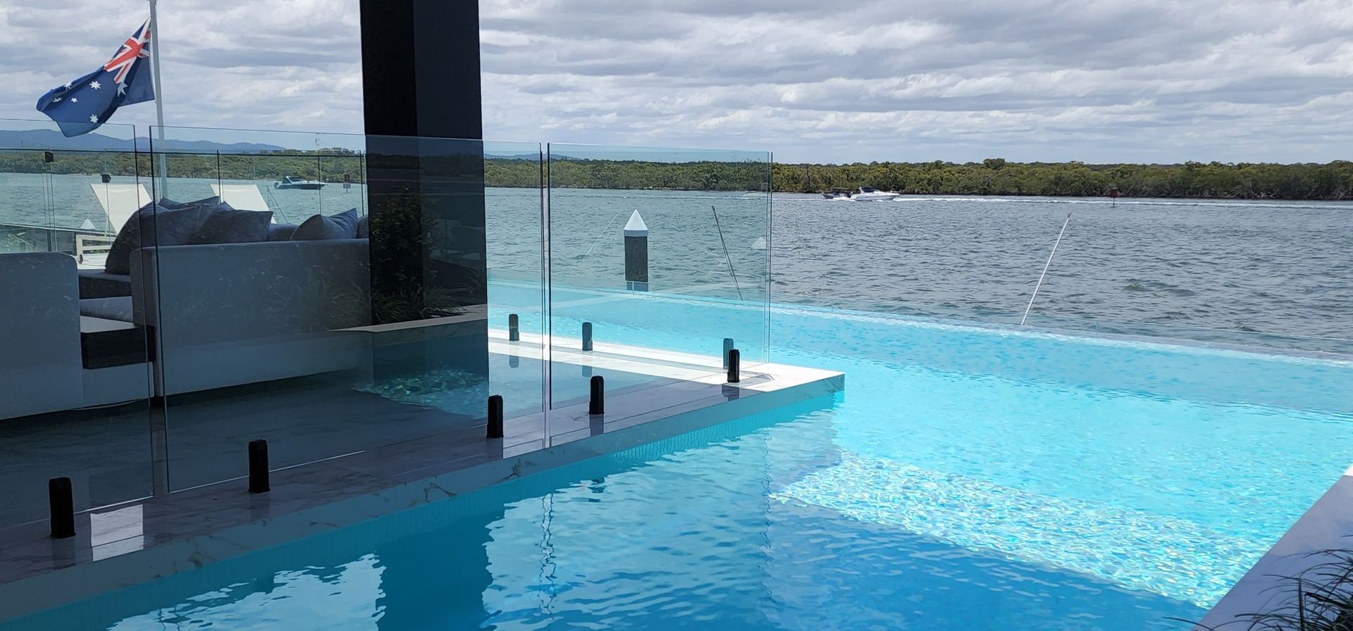 An infinity pool with glass walls overlooks a body of water and a distant treeline, with a flag waving in the sky.