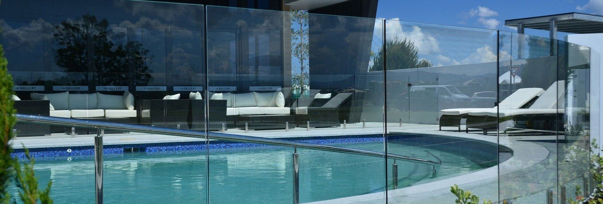 A pool with clear glass barriers, lounge chairs, and cushioned seating area on a patio.