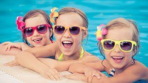 Three smiling girls in a pool wearing sunglasses and flower hair clips.
