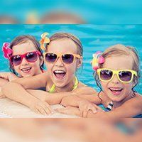 Three smiling girls in a pool, wearing sunglasses and flowers in their hair.