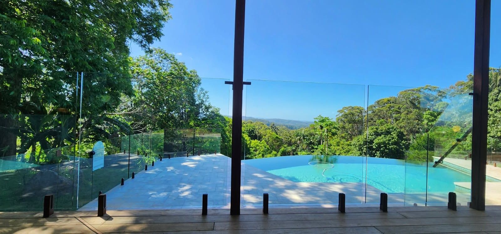 View of a pool and lush greenery from inside a building, framed by glass and wooden posts, on a sunny day.