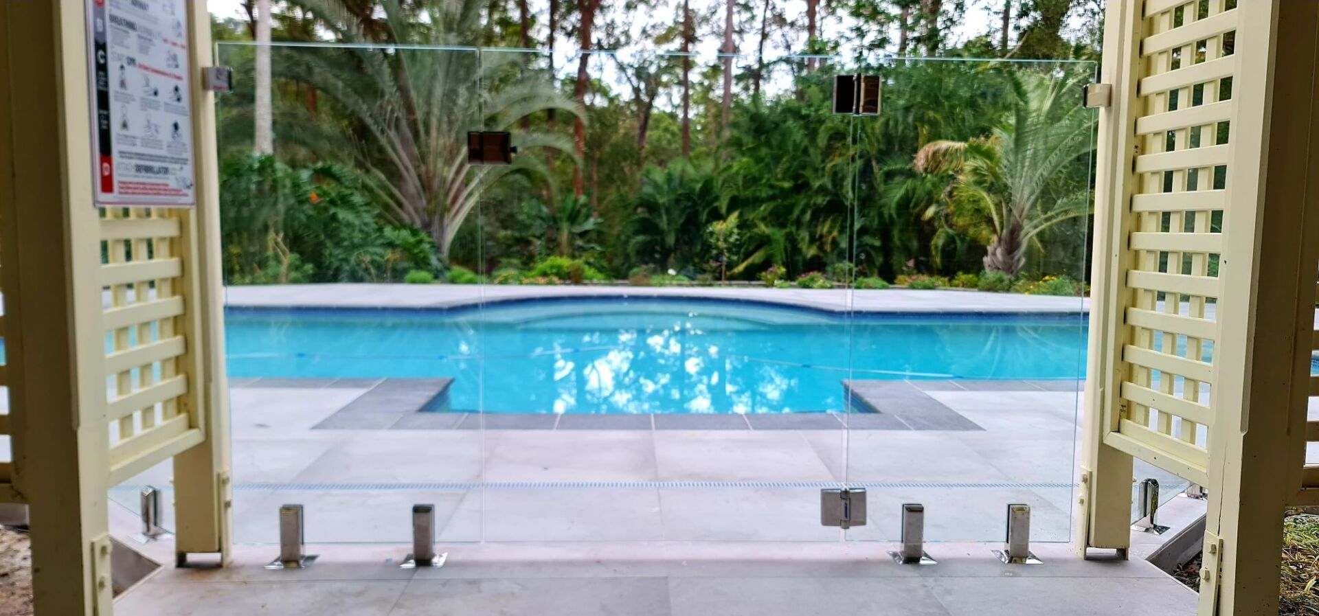 View of a pool through a glass fence, framed by a gazebo, with lush greenery in the background.