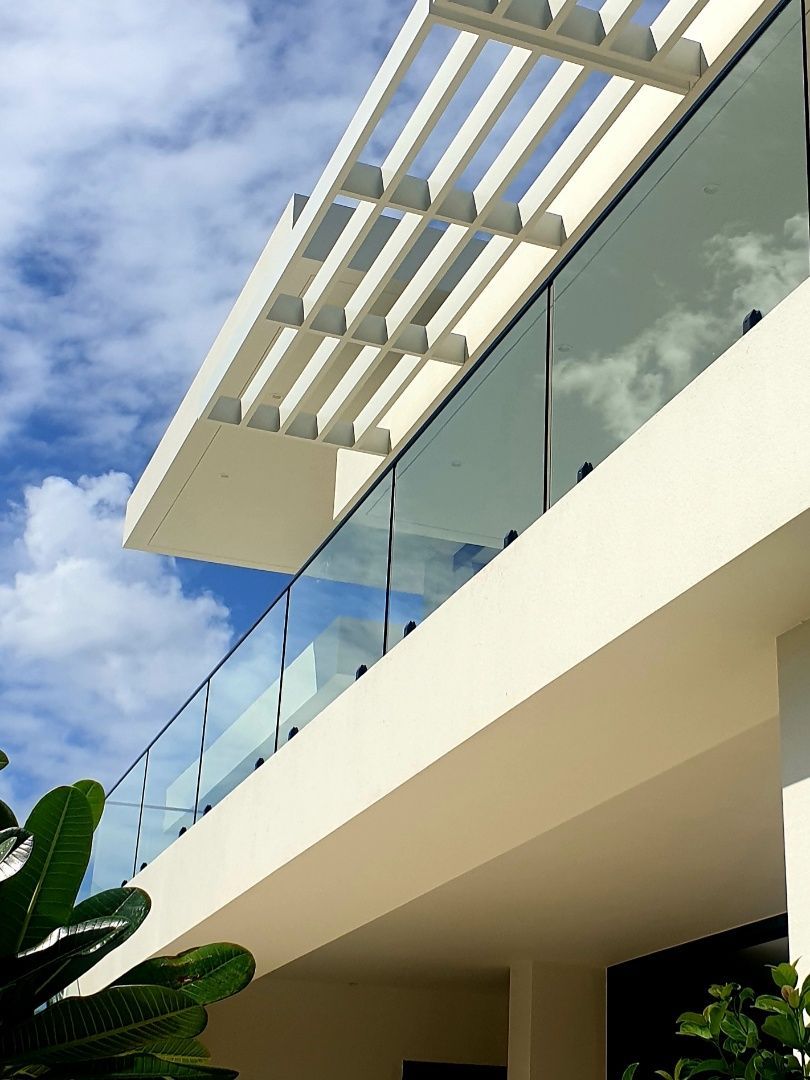 Modern building with white walls, glass balcony, and a pergola against a blue sky with clouds.