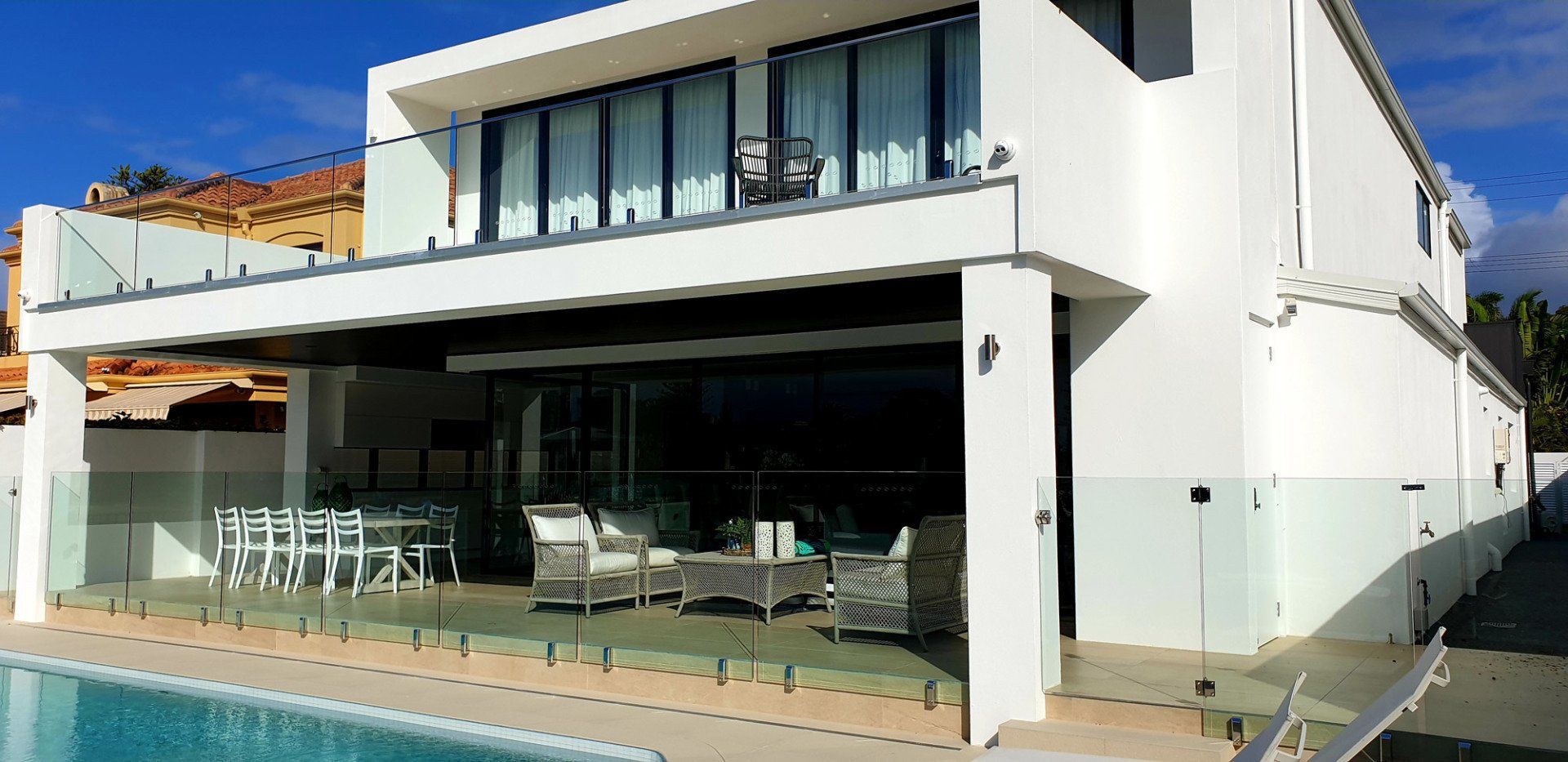 Modern white house with a pool, glass railings, and patio furniture. Bright blue sky.
