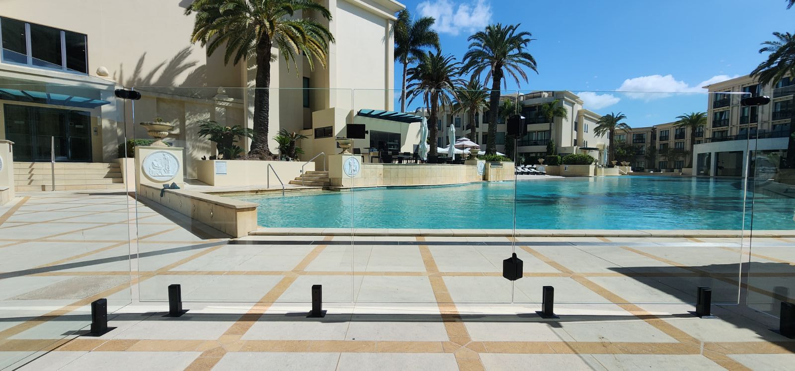 A large outdoor swimming pool with beige tiles, palm trees, and buildings under a blue sky.