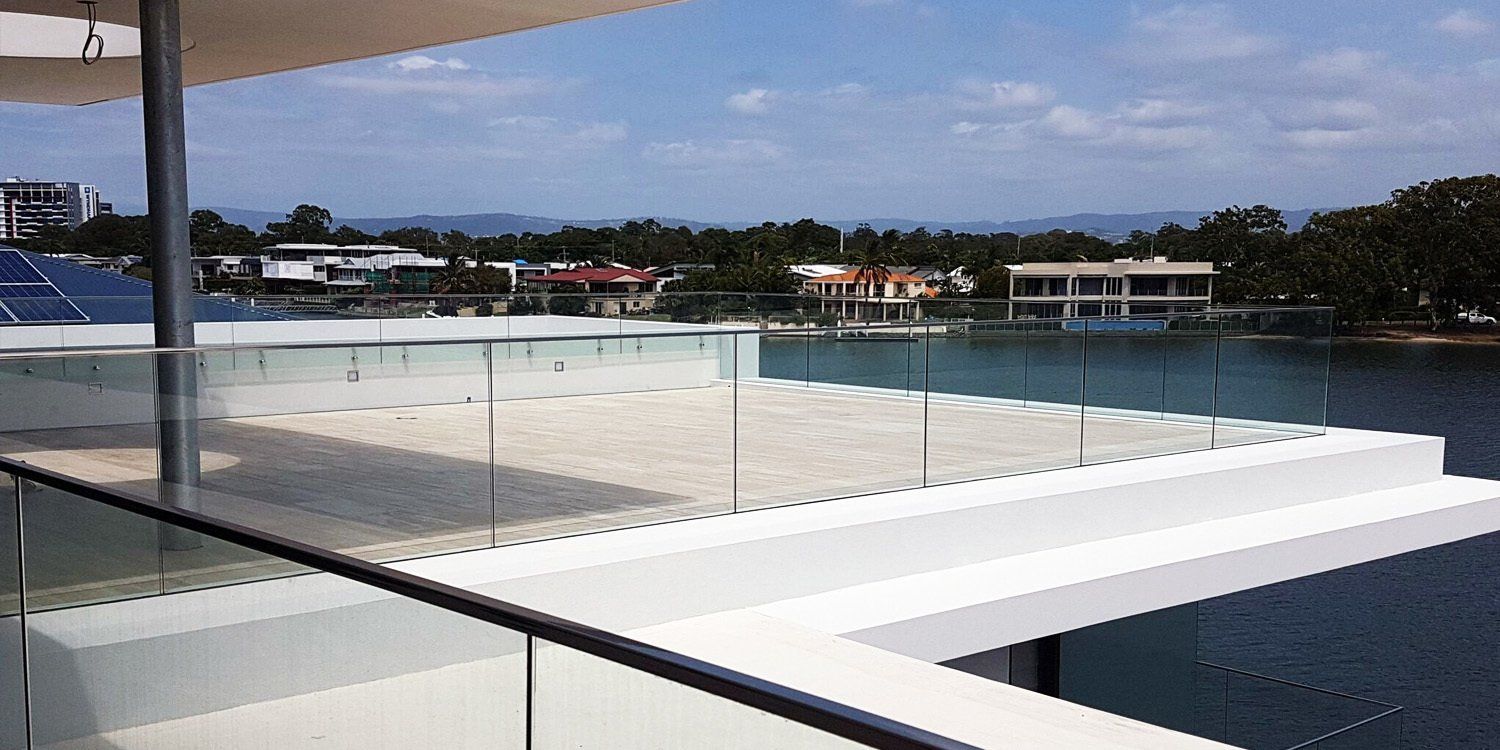 Modern waterfront home with glass railings, overlooking the water and other houses on a sunny day.