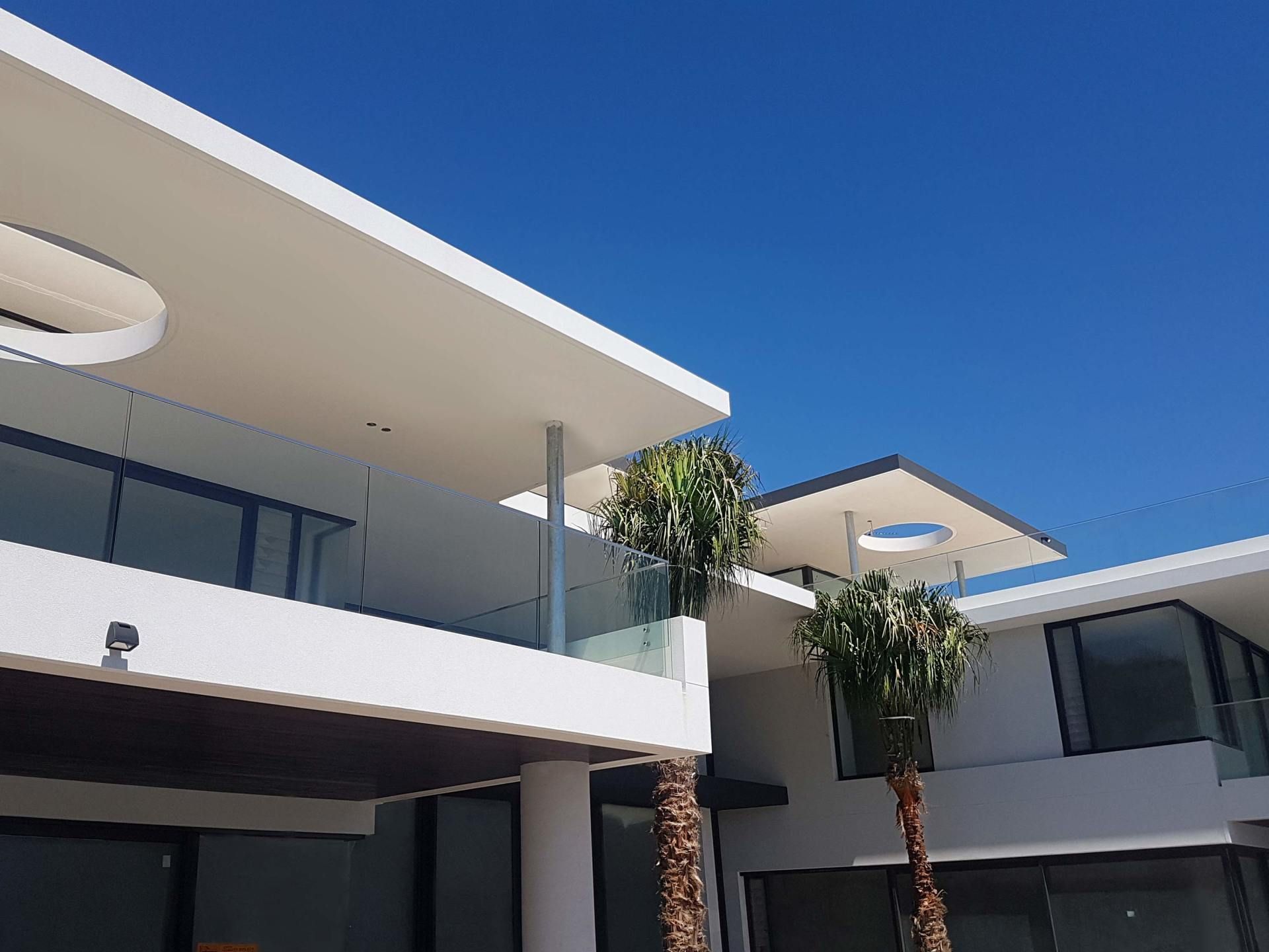 Modern white building with clear glass balconies and palm trees against a bright blue sky.