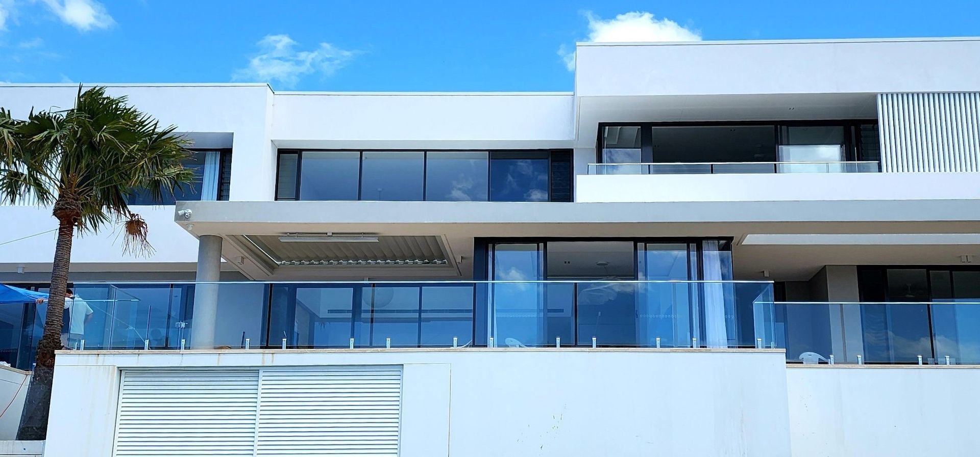 Modern white building with large windows, glass balconies, and a palm tree against a blue sky.
