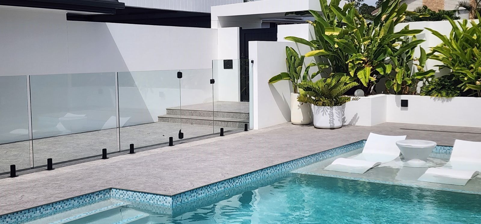 Outdoor pool with white lounge chairs and greenery, enclosed by a glass fence.