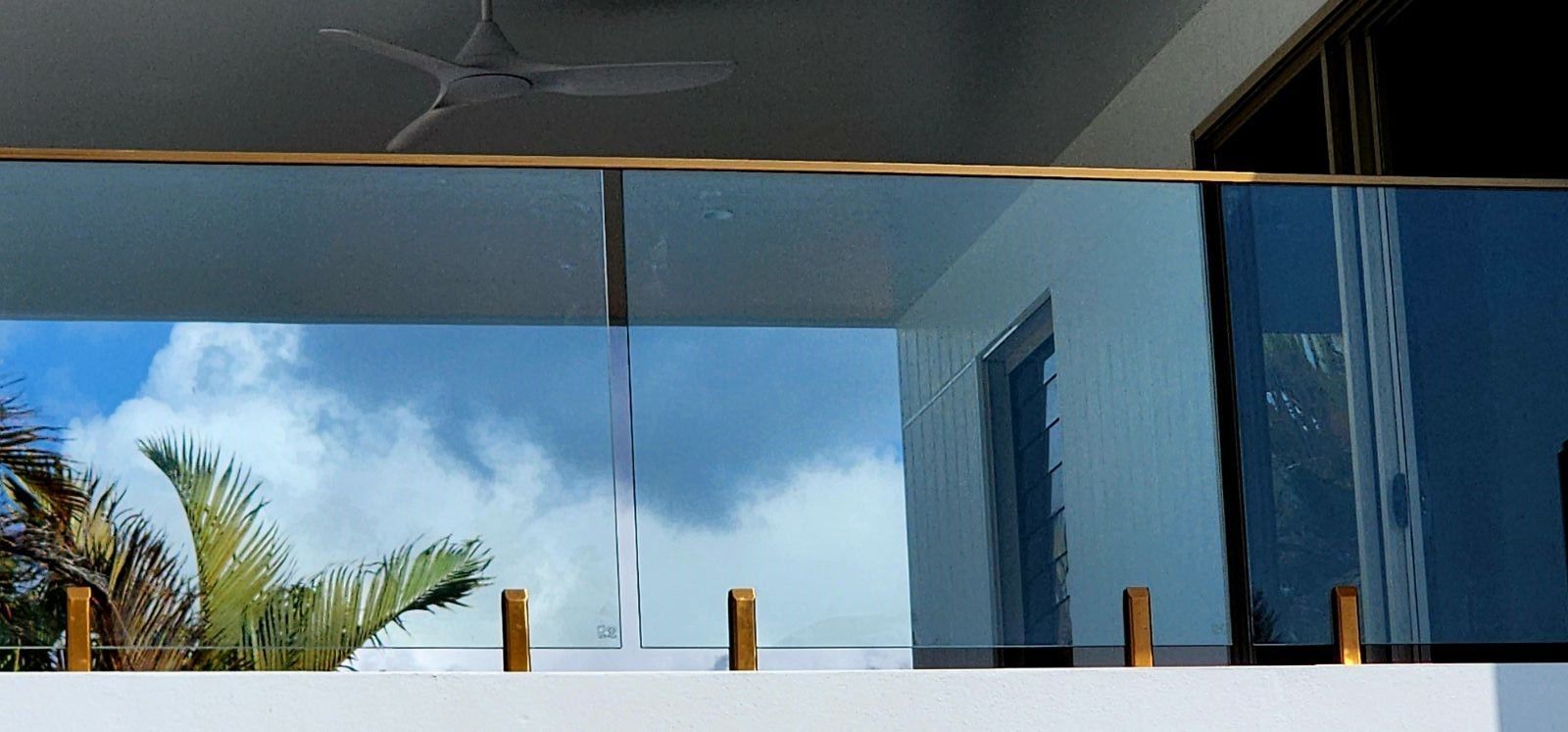 Glass balcony railing reflecting clouds and a palm tree; sunny day.