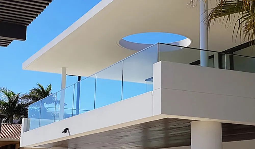 Modern white building with glass balcony overlooking palm trees under blue sky.