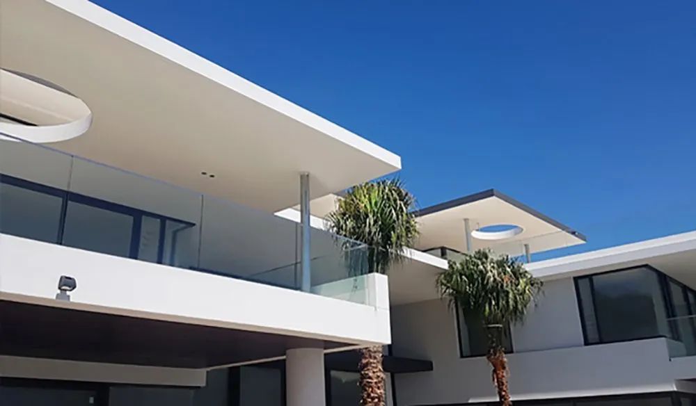 Modern white villa with glass railings and palm trees against a clear blue sky.
