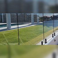 Glass fence alongside a grassy lawn, with white columns and a pool visible.