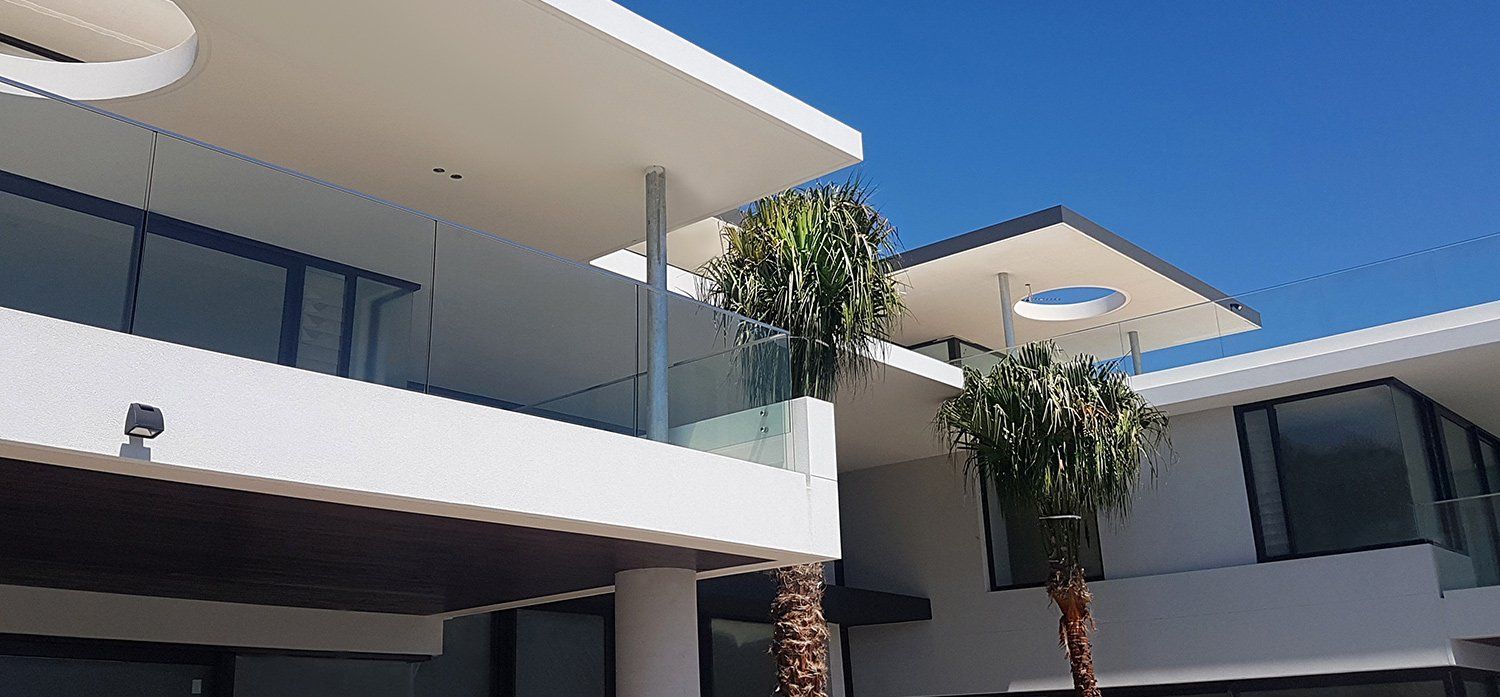 Modern white building with glass balconies and palm trees against a bright blue sky.