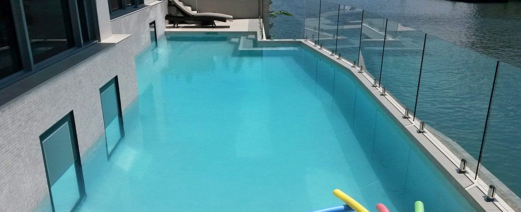 A rectangular swimming pool next to a building, water is bright blue, pool noodles float in the water.